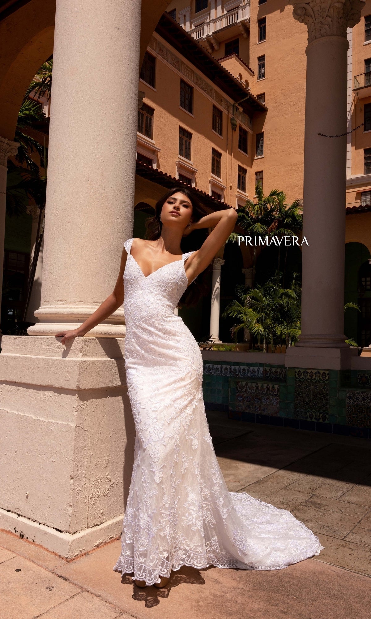 A woman models the Long Wedding Dress 11107 by Primavera, featuring an ivory hue and delicate v-neckline, as she poses beside a column outside a Mediterranean-style building with